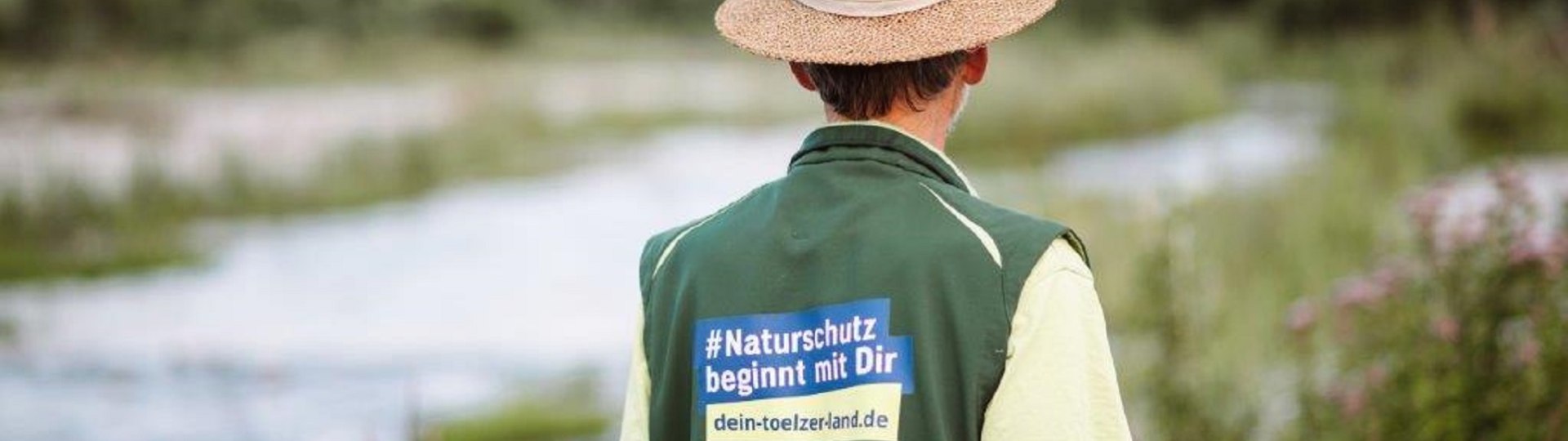 Ein Isarranger mit Strohhut und grüner Weste mit dem Rückenaufdruck "Naturschutz beginnt mit Dir", © Landratsamt Bad Tölz-Wolfratshausen, Foto Leonie Lorenz Ein Isarranger mit Strohhut und grüner Weste mit dem Rückenaufdruck "Naturschutz beginnt mit Dir", © Landratsamt Bad Tölz-Wolfratshausen, Foto Leonie Lorenz