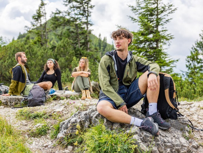 Das Bild zeigt vier junge Erwachsene, die auf einem Berg rasten,. Sie sitzen zwischen Felsen und Rucksäcken, umgeben von grünen Bäumen und bewölktem Himmel., © Tölzer Land Tourismus|farmhouse 7 Das Bild zeigt vier junge Erwachsene, die auf einem Berg rasten,. Sie sitzen zwischen Felsen und Rucksäcken, umgeben von grünen Bäumen und bewölktem Himmel., © Tölzer Land Tourismus|farmhouse 7