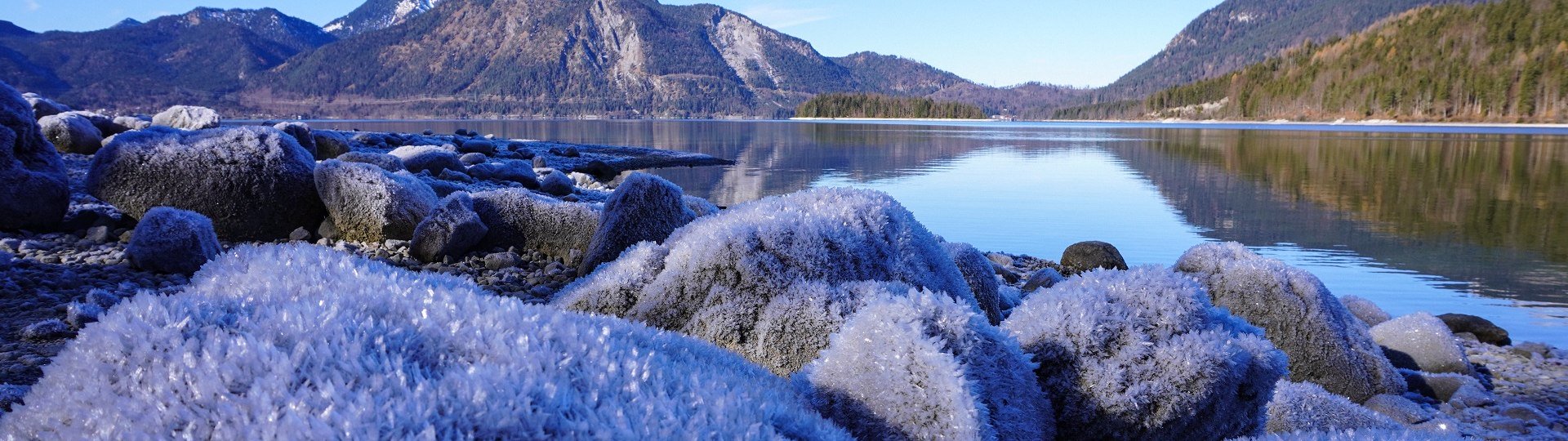Raureif liegt auf den großen Steinen am Walchenseeufer. Über dem glatten See spiegelt sich die Berglandschaft, © Landratsamt Bad Tölz-Wolfratshausen|Foto: A. Fischer Raureif liegt auf den großen Steinen am Walchenseeufer. Über dem glatten See spiegelt sich die Berglandschaft, © Landratsamt Bad Tölz-Wolfratshausen|Foto: A. Fischer