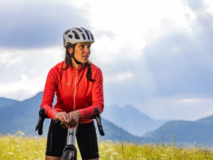 Bitte nutze die ausgewiesenen Wege.
Das Bild zeigt: Eine Frau mit Fahrradhelm und roter Sportjacke lehnt an ihrem Fahrrad, blickt nach rechts. Sie steht auf einer Blumenwiese vor einer Bergkulisse. Sonnenstrahlen brechen durch die Wolken am Himmel und beleuchten die Szene stimmungsvoll., © Tölzer Land Tourismus|farmhouse7