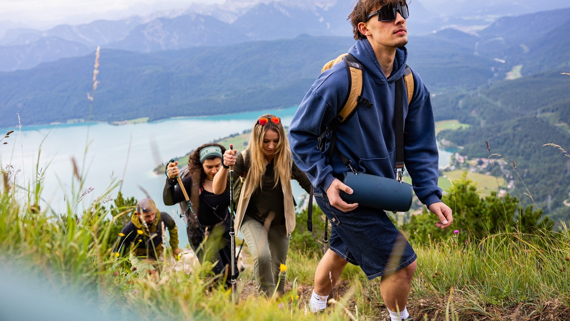 Das Bild zeigt: F&uuml;nf junge Menschen wandern einen grasbewachsenen Berg hinauf, mit Wanderst&ouml;cken und Rucks&auml;cken. Im Vordergrund tr&auml;gt ein Mann in blauer Kapuzenjacke eine tragbare Musikbox. Im Hintergrund der t&uuml;rkisfarbene Walchensee und bewaldete Berge unter bew&ouml;lktem Himmel., &copy; T&ouml;lzer Land Tourismusfarmhouse7