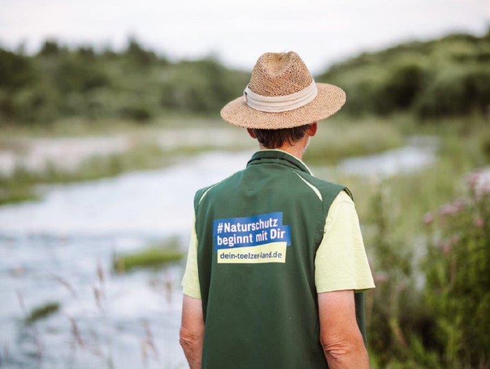 Ein Isarranger mit Strohhut und gr&uuml;ner Weste mit dem R&uuml;ckenaufdruck "Naturschutz beginnt mit Dir", &copy; Landratsamt Bad T&ouml;lz-Wolfratshausen, Foto Leonie Lorenz