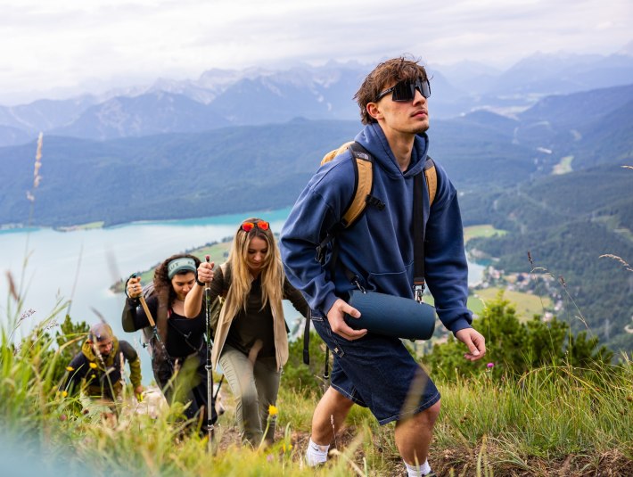 Das Bild zeigt: Fünf junge Menschen wandern einen grasbewachsenen Berg hinauf, mit Wanderstöcken und Rucksäcken. Im Vordergrund trägt ein Mann in blauer Kapuzenjacke eine tragbare Musikbox. Im Hintergrund der türkisfarbene Walchensee und bewaldete Berge unter bewölktem Himmel., © Tölzer Land Tourismusfarmhouse7