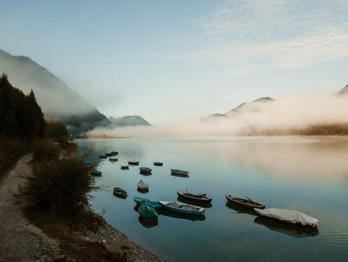 Eine ruhige Wasserlandschaft mit mehreren Booten, die am Ufer und auf dem Wasser liegen. Im Hintergrund sind Berge und eine dünne Nebelschicht zu sehen, unter einem bewölkten Himmel., © Tölzer Land Tourismus|Leonie Hartung
