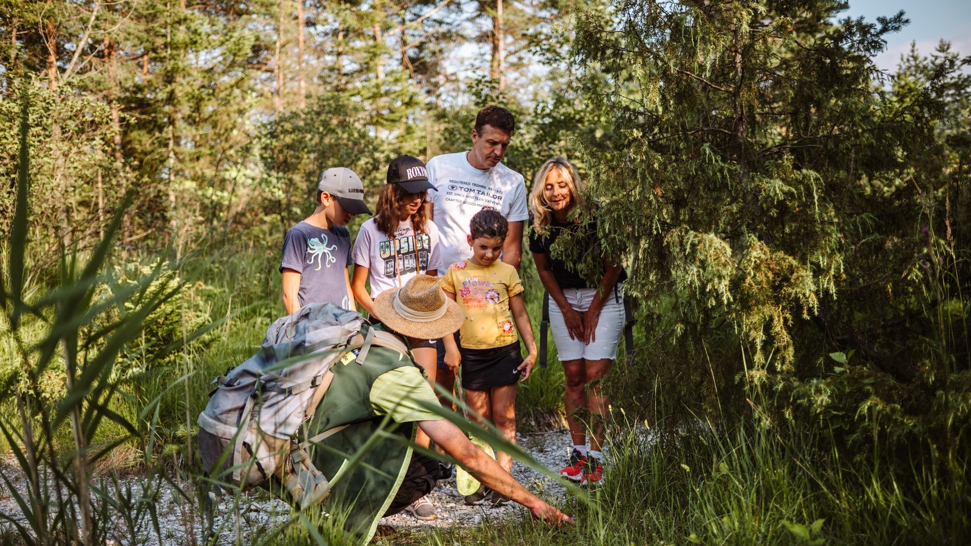 Eine 5-köpfige Familie schaut in einem Waldgebiet interessiert auf einen Naturschutzranger in grüner Kleidung mit Strohhut, der auf etwas im Gras zeigt., © Tölzer Land Tourismus|Leonie Lorenz