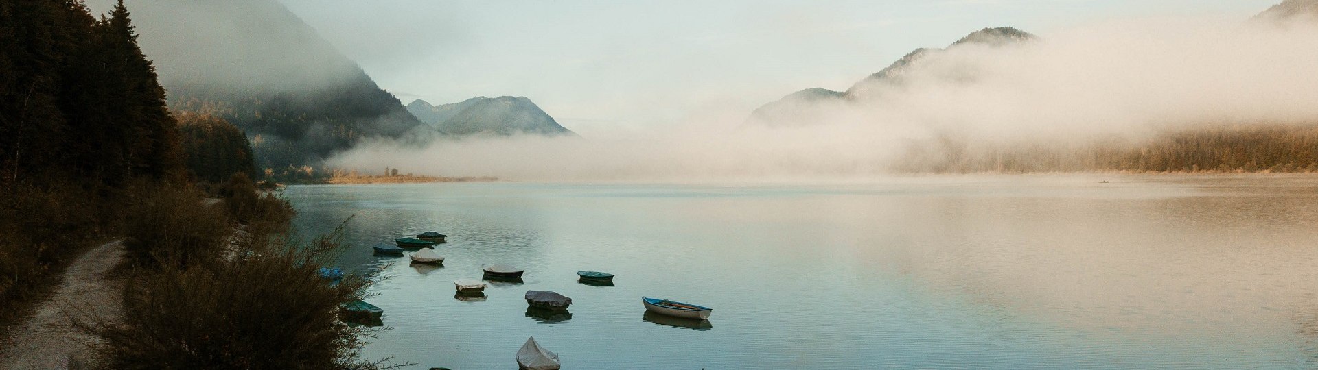 Eine ruhige Wasserlandschaft mit mehreren Booten, die am Ufer und auf dem Wasser liegen. Im Hintergrund sind Berge und eine dünne Nebelschicht zu sehen, unter einem bewölkten Himmel., © Tölzer Land Tourismus|Leonie Hartung