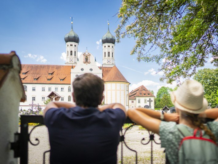 Das Bild zeigt ein  sommerlich gekleidetes Paar von Hinten, das einem schmiedeeisernen Gel&auml;nder gelehnt die pr&auml;chtige barocke Basilika des Klosters Benediktbeuern mit seinen zwei markanten Zwiebelt&uuml;rmen betrachtet., &copy; T&ouml;lzer Land Tourismus, Dietmar Denger